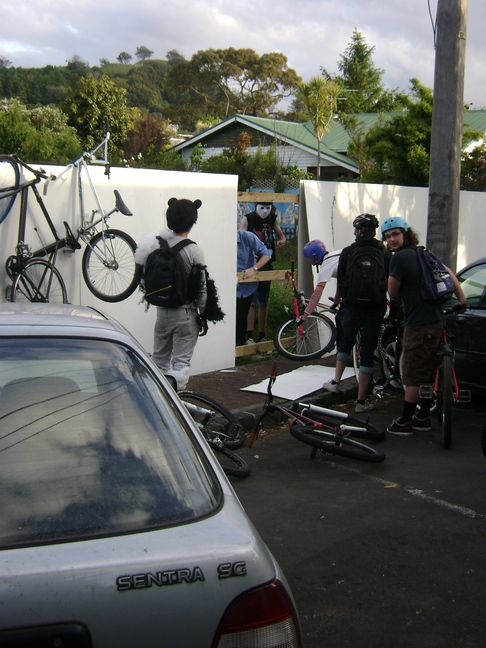 Rear view of cyclists standing with their bikes, busting through a fence panel with a house and trees behind it